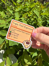 Stardew Valley Triple Shot Espresso pixel art item card die-cut sticker with espresso cup icon, held in hand outdoors against green leaves