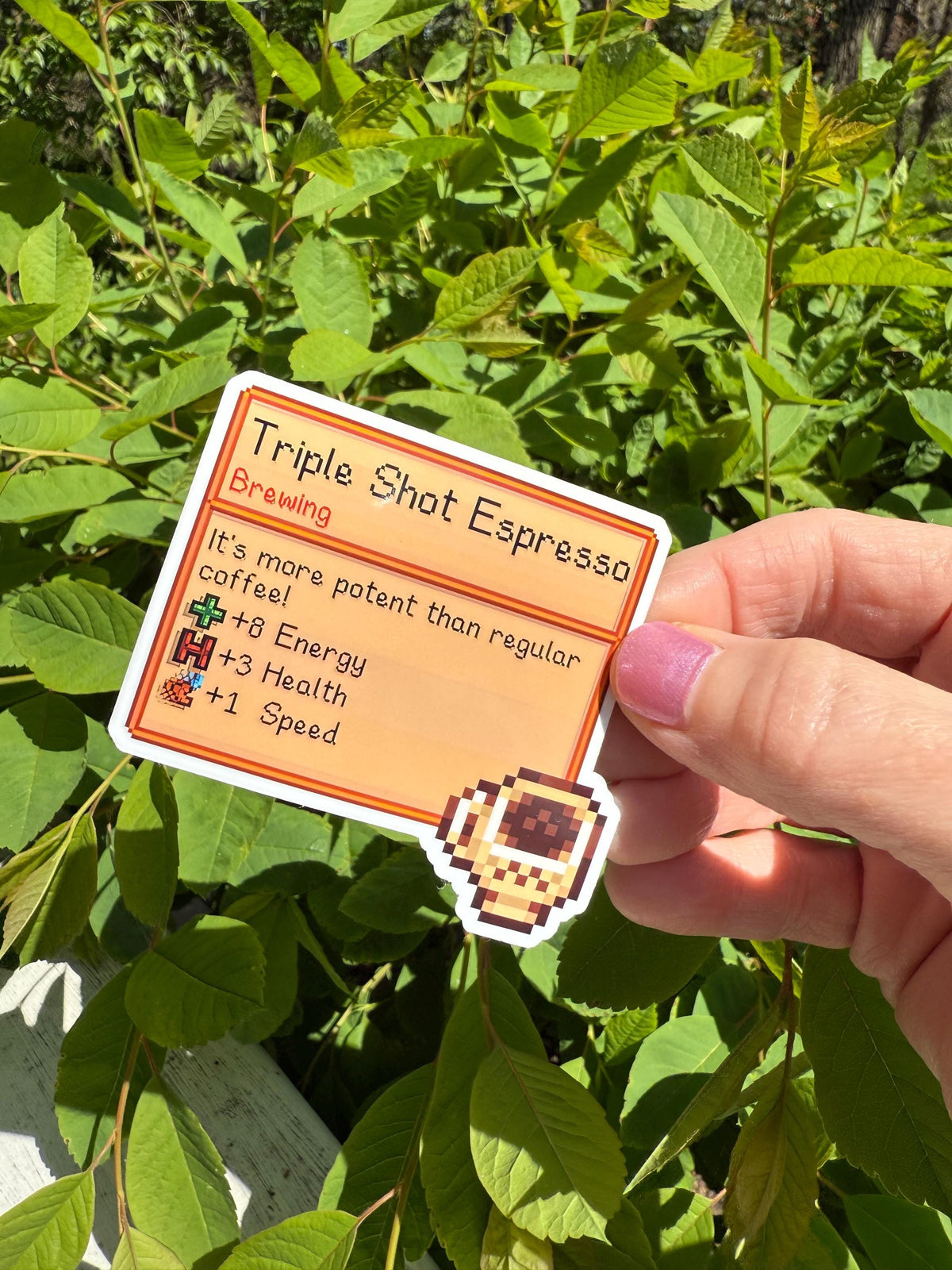 Stardew Valley Triple Shot Espresso pixel art item card die-cut sticker with espresso cup icon, held in hand outdoors against green leaves