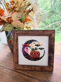Framed Halloween Witch Moon cross stitch in a dark brown frame, styled with a fall floral arrangement