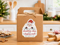 Christmas shopping small sticker with a smiling Santa on a kraft shipping box, gingerbread cookies and holiday greenery in a cozy kitchen